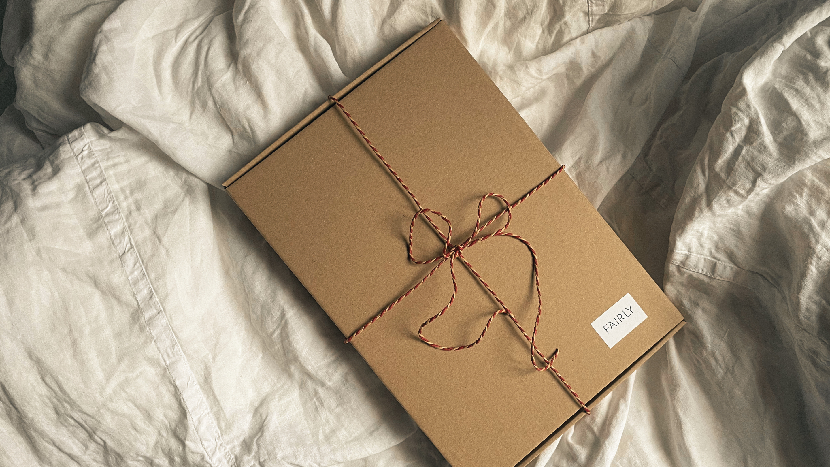 Ett fyrkantigt paket i brunt kraftpapper, med ett jutesnöre i rött och vitt knutet i en rosett och en liten vit etikett med FAIRLY skrivet i högra hörnet, ligger på ett vitt påslakan som veckar sig mjukt