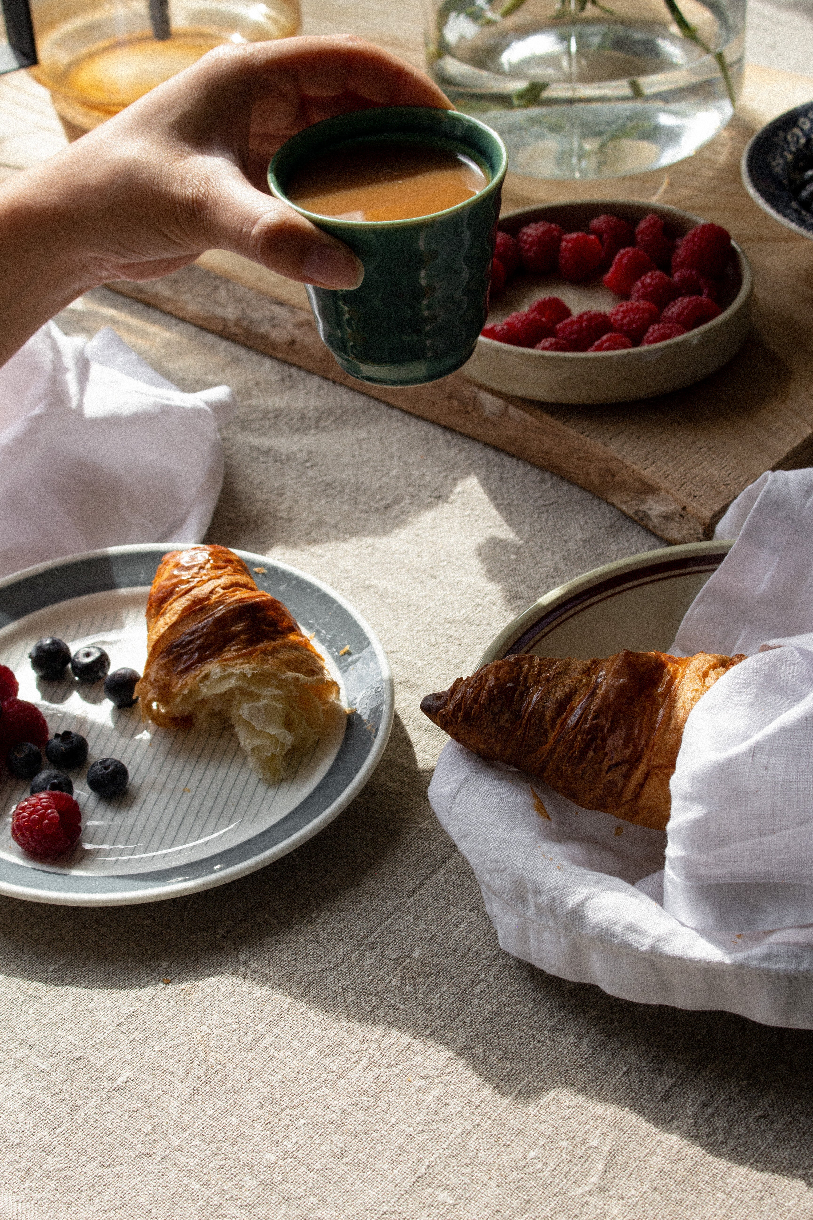 Bilden föreställer ett dukat bord med vita linneservetter, croissanter och bär. En hand håller en kaffekopp i mitten av bilden. 