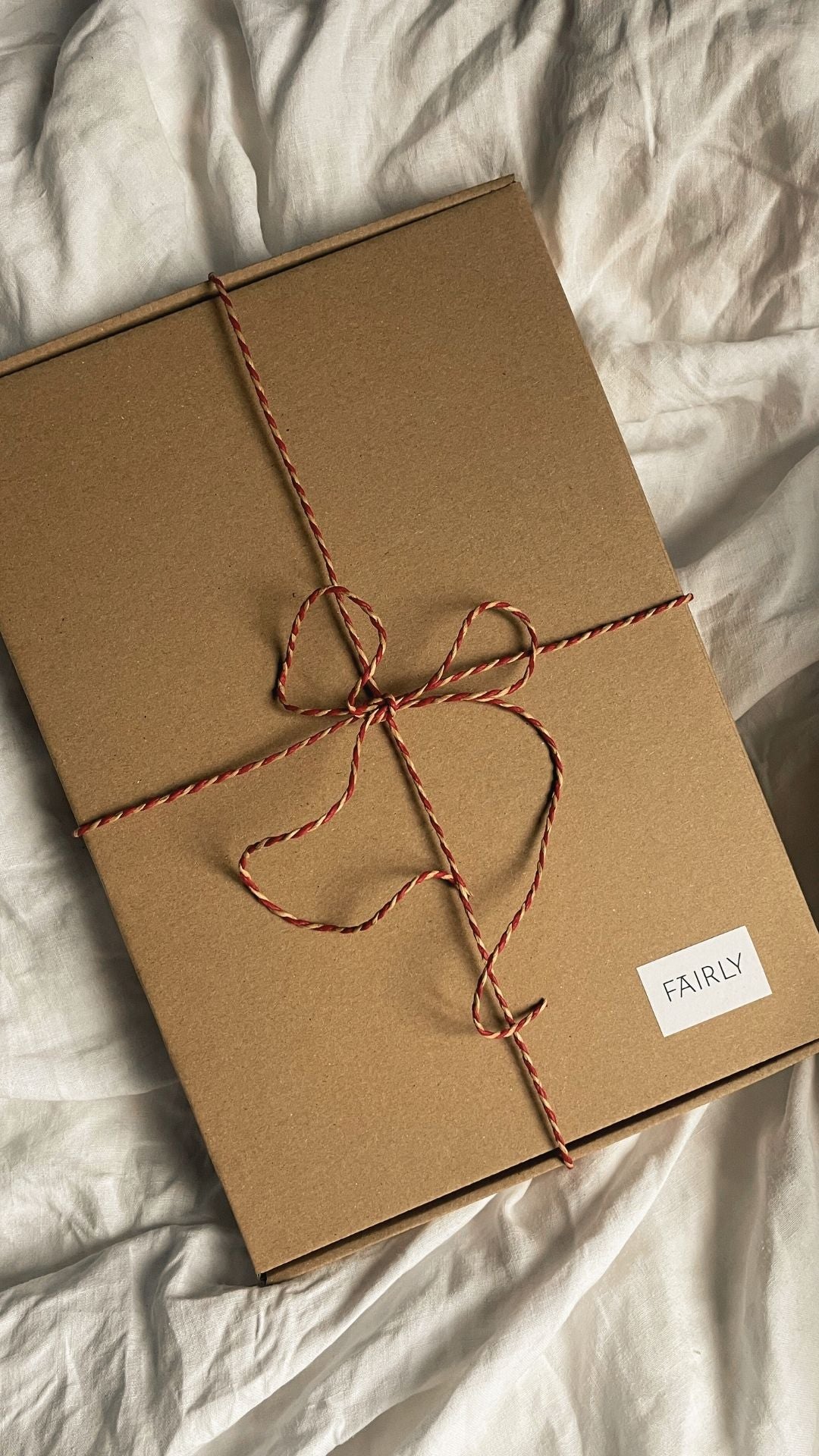 Ett fyrkantigt paket i brunt kraftpapper, med ett jutesnöre i rött och vitt knutet i en rosett och en liten vit etikett med FAIRLY skrivet i högra hörnet, ligger på ett vitt påslakan som veckar sig mjukt